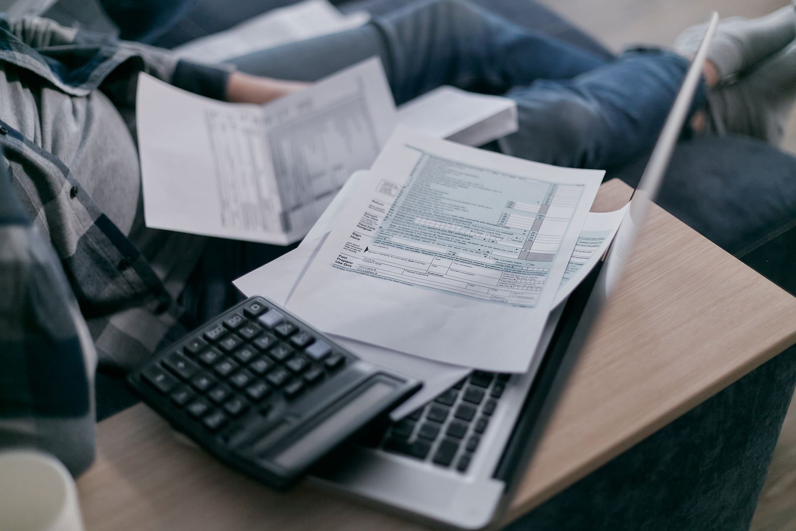 Formulieren, een rekenmachine en een laptop op schoot: iemand werkt thuis de administratie bij.